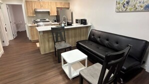 Un espacio habitable compacto con kitchenette, un sofá de cuero negro, una mesita blanca pequeña y una silla.