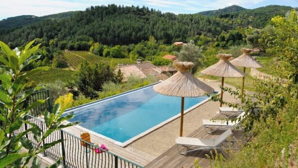 A rectangular swimming pool with a wooden deck, surrounded by lounge chairs and thatched umbrellas, set against a backdrop of lush green hills and a clear sky. - Le mas de Jeanne (Rocher)