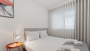 Ett hotellrum med en säng, ett nattduksbord med en lampa och ett fönster med tunna, genomskinliga gardiner.