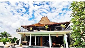 A traditional building with a tiled roof, a balcony with a railing, and a pool area with lounge chairs. - KAMAR MERAH. Comfortable 1BR Beachfront Apartment With AC, Pool & Kitchenette. (Tegallengah)