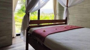 A bedroom with a wooden bed, white curtains, and a view of greenery outside. - KAMAR MERAH. Comfortable 1BR Beachfront Apartment With AC, Pool & Kitchenette. (Tegallengah)