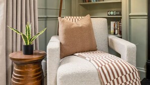 Un salón acogedor con un sillón de color claro, una manta estampada, una lámpara de pie alta, una pequeña mesita de madera con una planta en maceta y una estantería al fondo.