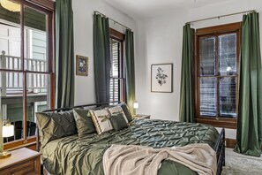 Un lit bien fait, avec une couverture verte et des oreillers, une table de chevet avec une lampe, et une fenêtre ornée de rideaux verts.