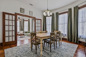 Une salle à manger avec une table et des chaises en bois, un lustre et de grandes fenêtres avec des rideaux.