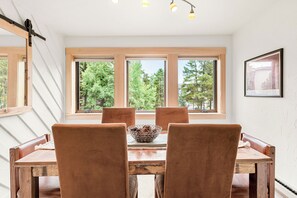 Un coin repas avec une table en bois et quatre chaises, de grandes fenêtres donnant sur la forêt et un tableau encadré au mur.