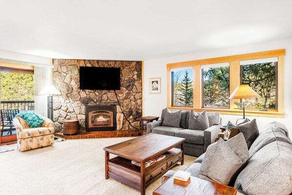 A living room with a stone fireplace, a television mounted on the wall, and a large window with a view of trees. - Corner Unit in the Center of Town, Walk to Lifts (Breckenridge)