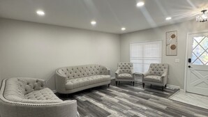 A room with a grey tufted sofa, two armchairs, a wooden herringbone floor, recessed ceiling lights, and a white door with a window. - Luxury House Perfect location-3BR Sterling Heights  (Sterling Heights)