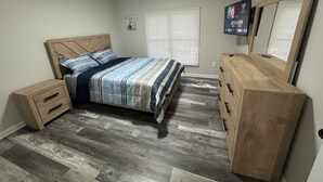 A bedroom with a wooden bed, a dresser, a TV on the wall, and a window with blinds. - Luxury House Perfect location-3BR Sterling Heights  (Sterling Heights)