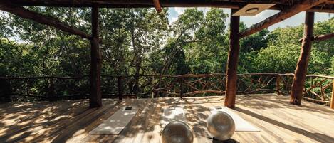 An outdoor wooden deck with exercise mats and yoga balls, surrounded by trees and a bamboo roof structure.