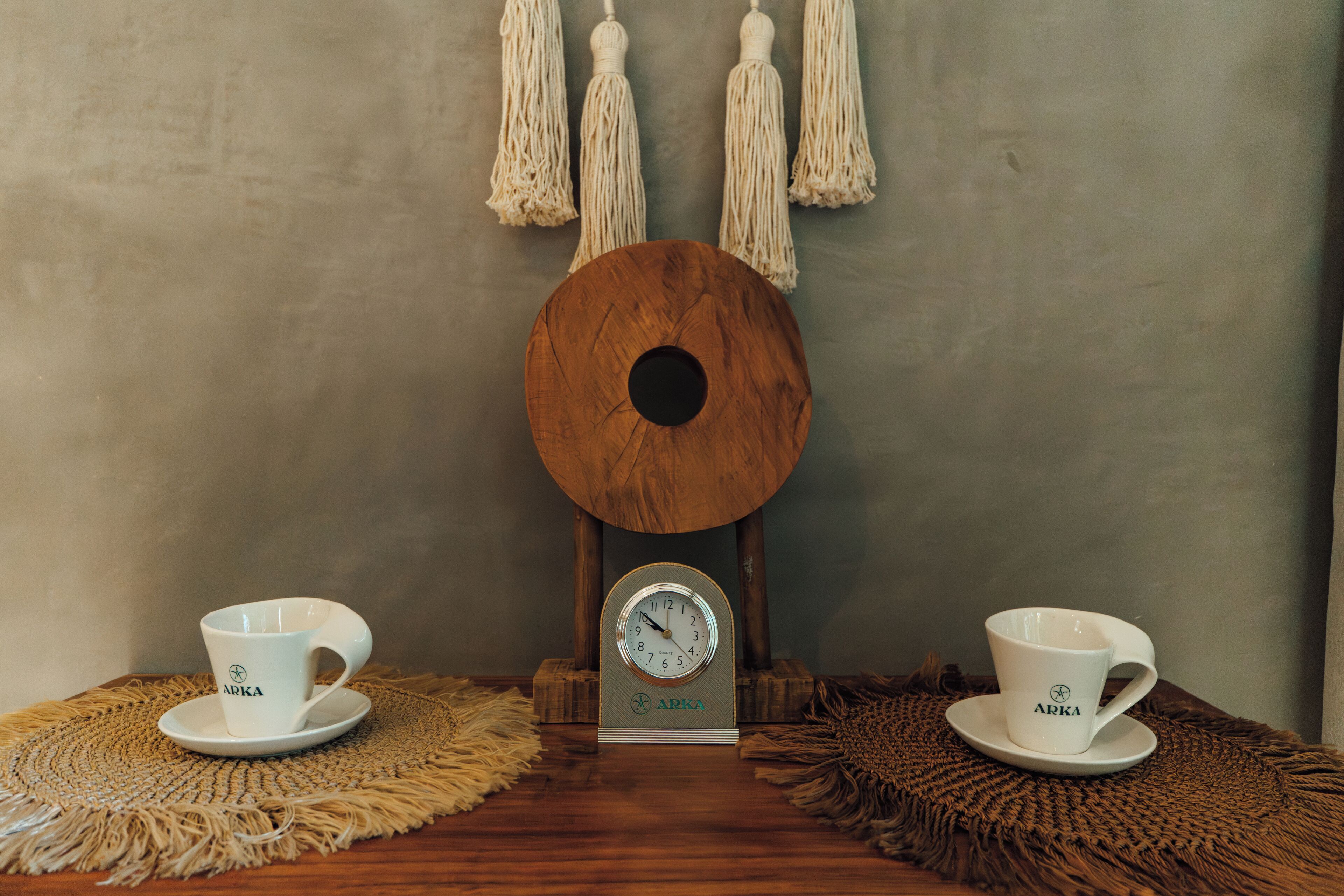 Deux tasses blanches, portant l’inscription « APKA », sont posées sur des sets de table en fibres tressées, sur une table en bois.