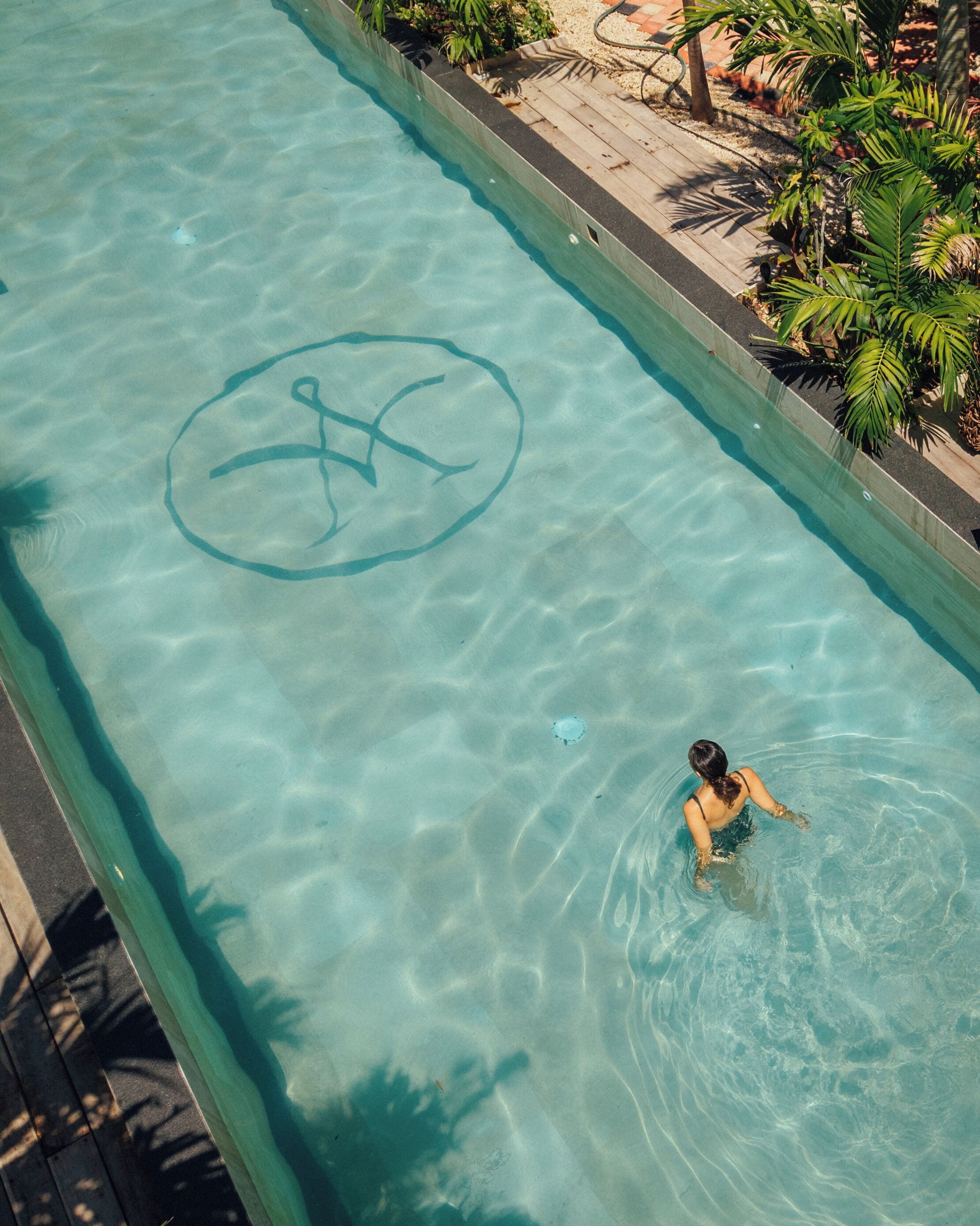 A swimming pool with a person swimming, surrounded by a wooden deck and palm trees.
