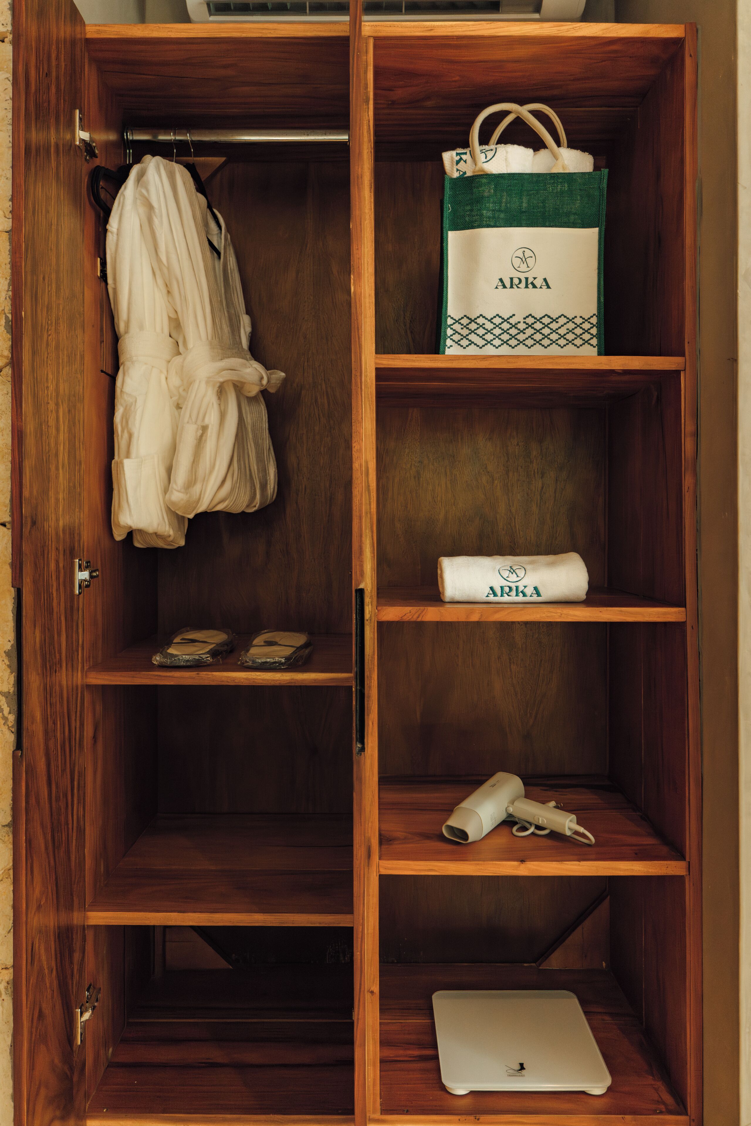 A wooden wardrobe with shelves containing a white robe, a green and white ARKA bag, a rolled-up ARKA item, a white laptop, and a white hairdryer.