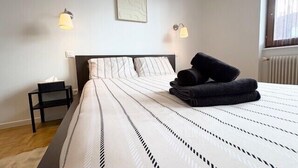 A neatly made bed with striped bedding, two bedside lamps, a small bedside table with a tissue box, and a window with blinds. - The Dutt's cocoon: spacious and charming T3 (Duttlenheim)