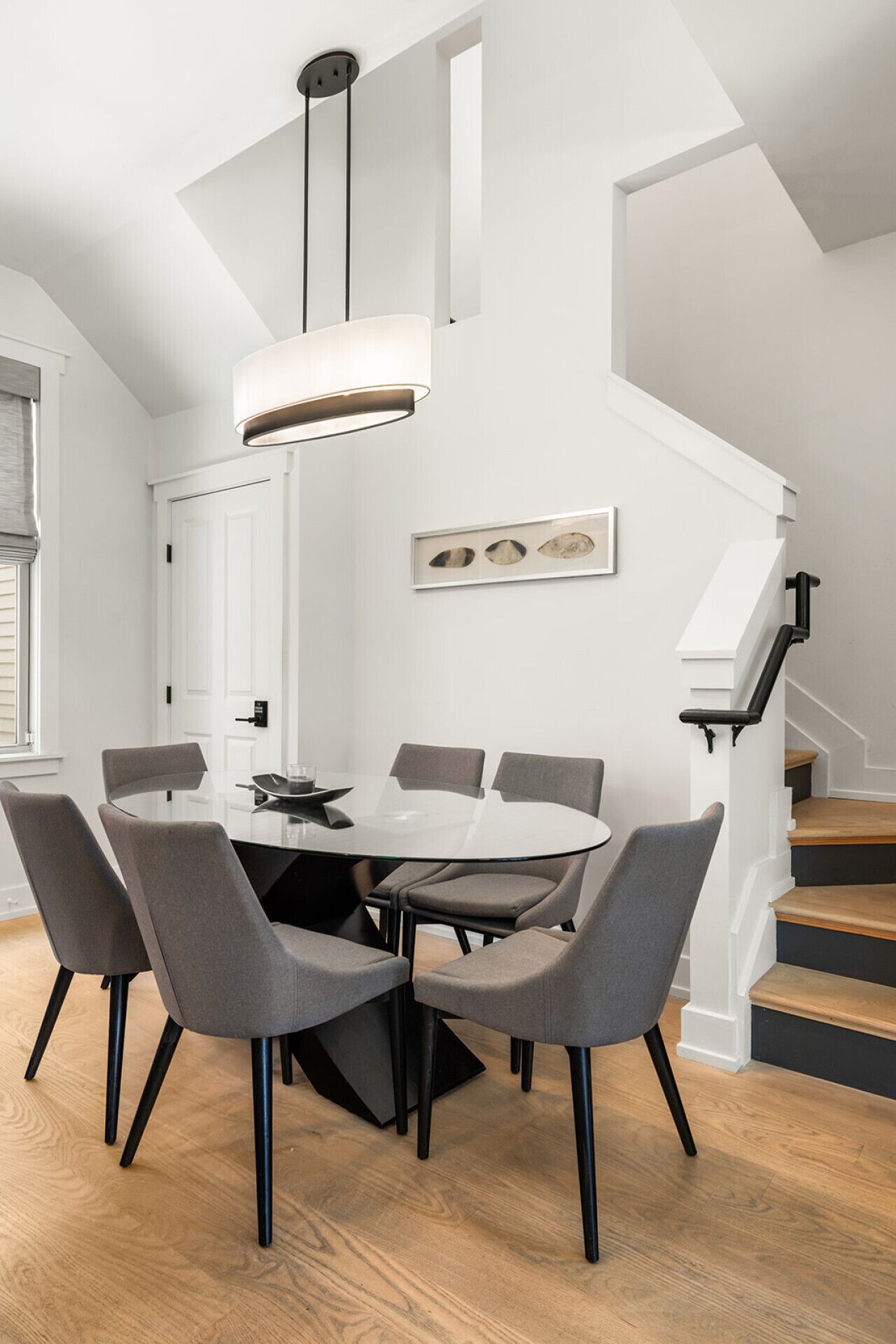 A modern dining area with a round glass table, six grey upholstered chairs, and a pendant light.