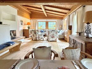 A cozy living room with wooden beams, floral-patterned sofas, a dining table with plates, and a fireplace.
