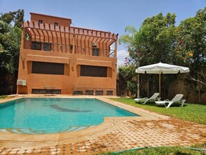 Un bâtiment à deux étages doté d’une piscine, d’un espace salon couvert et d’un jardin avec des arbres et des fleurs.