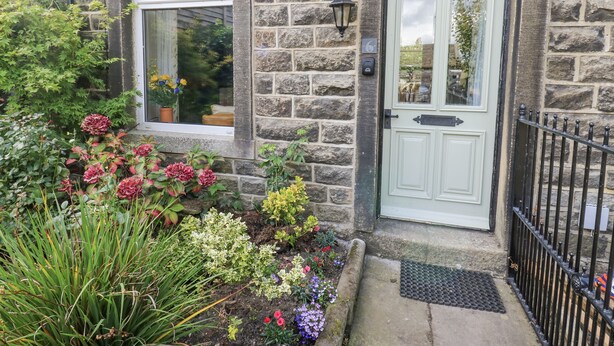 A stone building with a white door and window, surrounded by a garden with various plants and flowers. - Centenary Cottage (Skipton)
