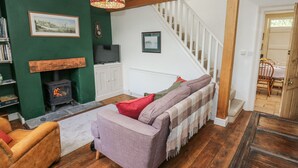 A cozy living room with a fireplace, a sofa, a wooden staircase, and a dining area. - Centenary Cottage (Skipton)