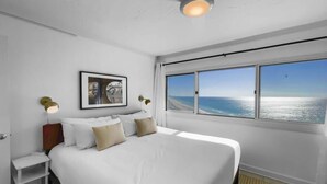 A modern hotel room with a large bed, a nightstand, a framed picture on the wall, and a window with a view of the sea.