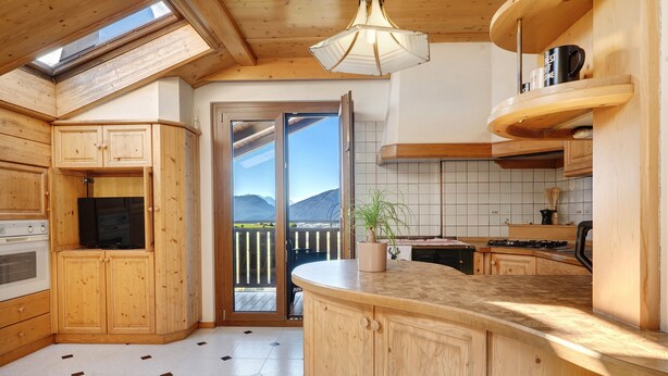 Une cuisine avec des meubles en bois, un îlot central, une plante en pot et une vue à travers la porte vitrée.
