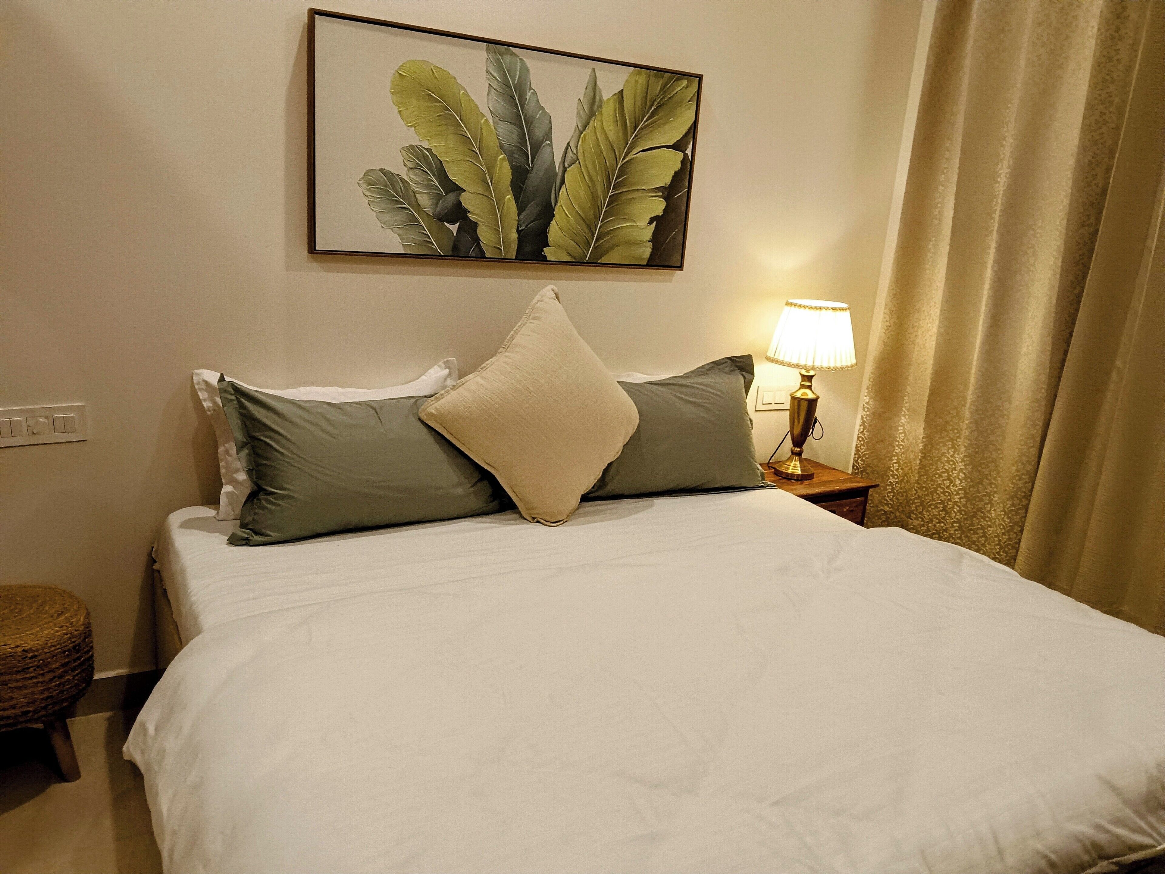 Una cama bien hecha con edredón blanco y dos almohadas, una planta en un marco en la pared, una lámpara de noche y una cortina.
