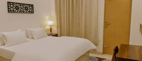 Un letto rifatto con lenzuola bianche, un comodino in legno con una lampada, una porta in legno e un piccolo tavolo con una sedia.