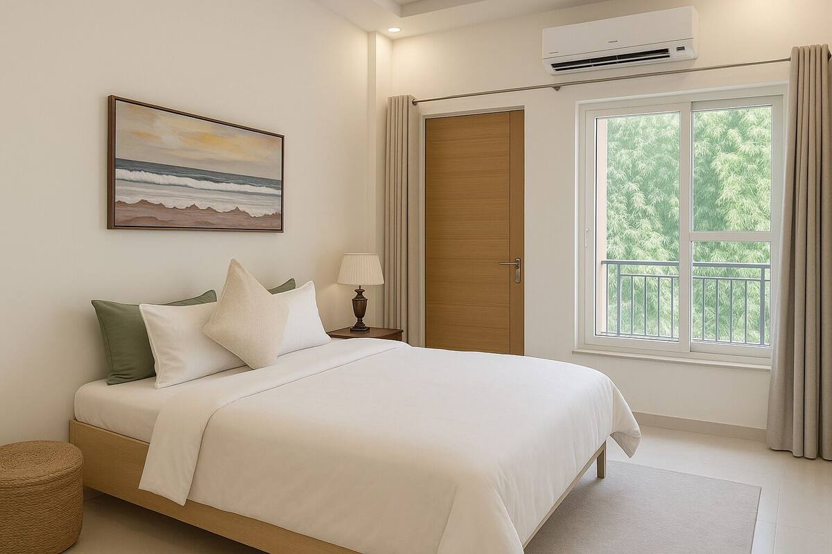 A bedroom with a bed, a nightstand, a lamp, a window with curtains, a door, and an air conditioning unit.