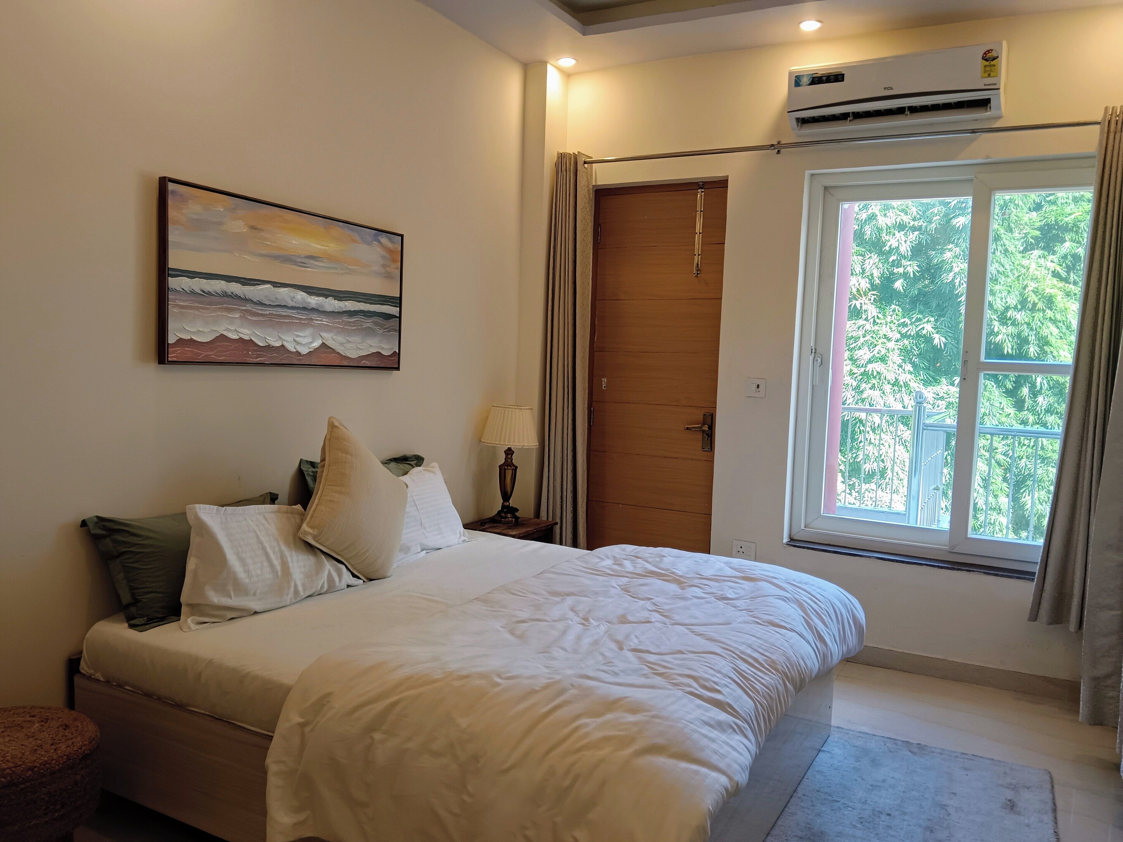 Um quarto com cama, um quadro do oceano, ar condicionado e janela com vista para árvores.