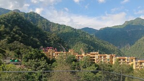 A hillside residential area with multiple yellow buildings, surrounded by lush green hills and trees. - Pennylane Hotel (Rishikesh)