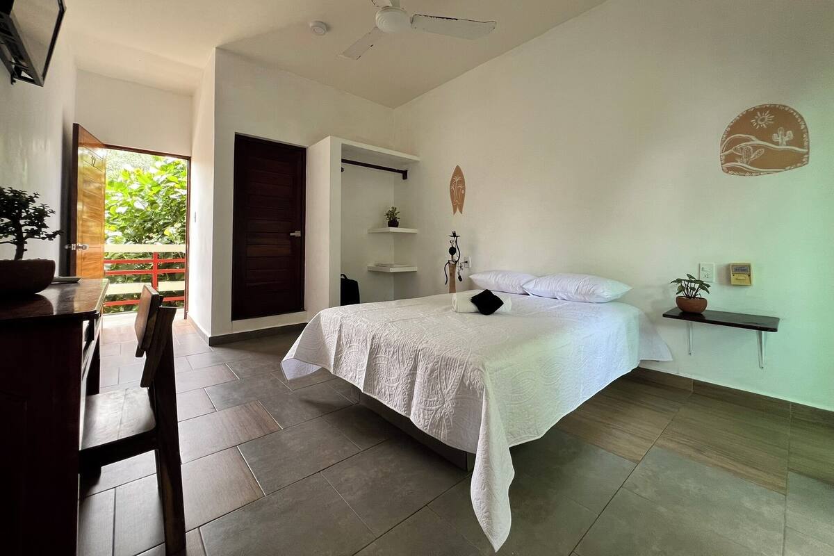A bedroom with a bed, a desk, a chair, a ceiling fan, and a view of a garden through an open door.