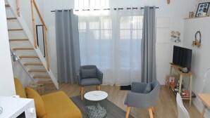 Un salon moderne avec un escalier, des fauteuils gris, une table basse blanche et un téléviseur posé sur un meuble en bois.