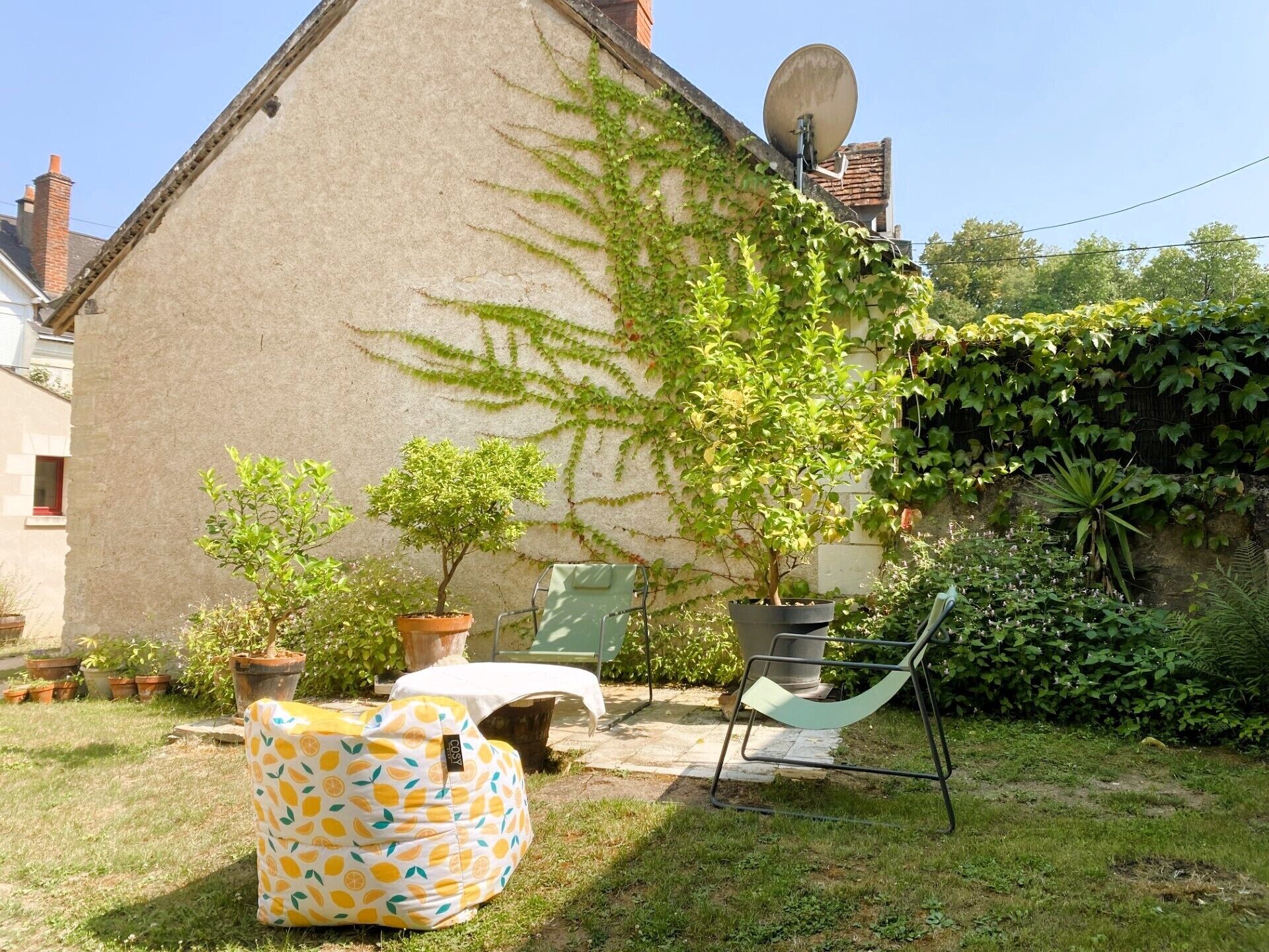 Ein Haus mit geneigtem Dach, einer Satellitenschüssel und einem Garten mit Kübelpflanzen und einer Hängematte.