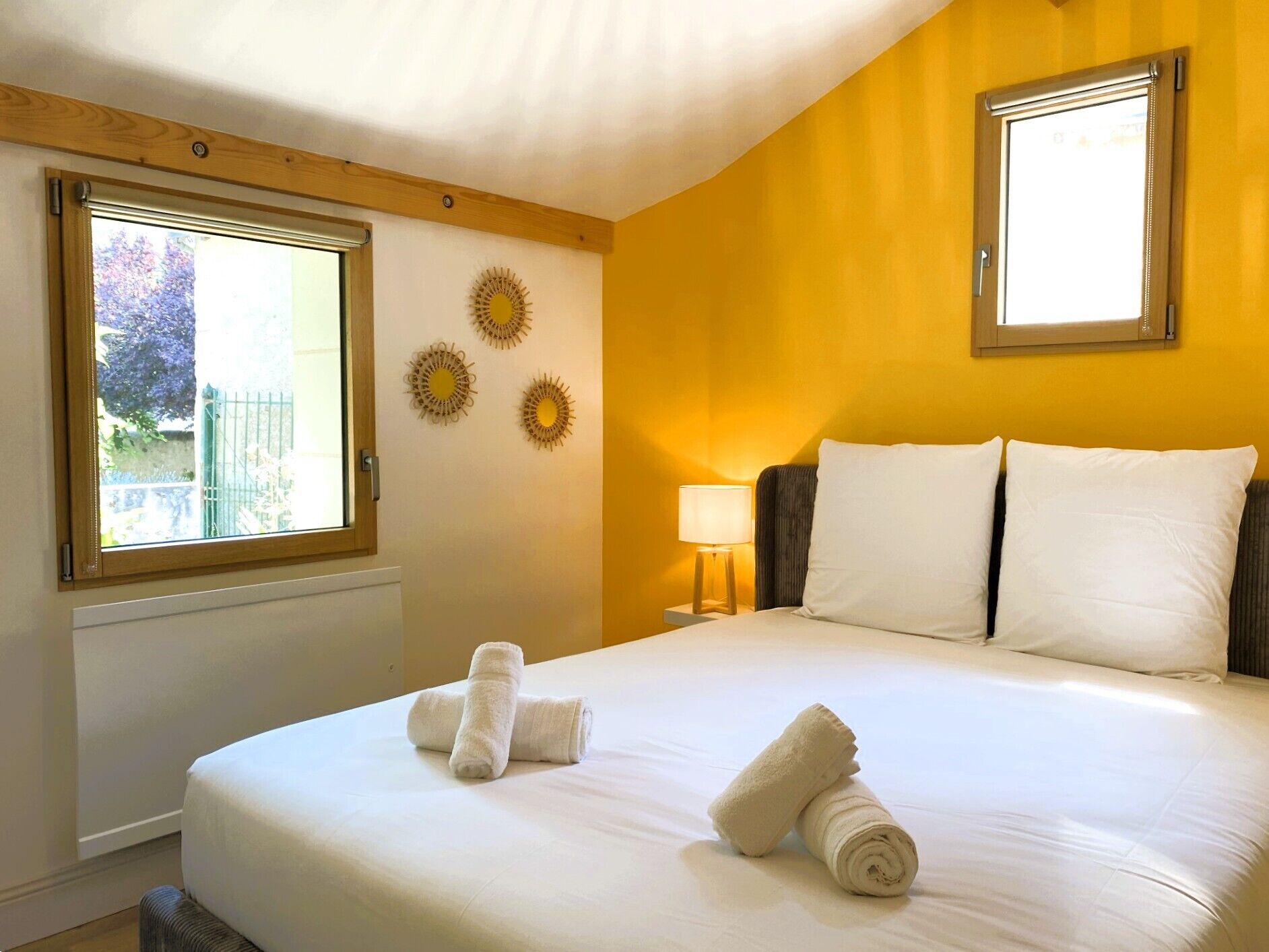 A hotel room with a large bed, two windows, a nightstand with a lamp, and decorative wall art.