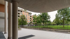 Un espacio al aire libre techado con suelo de baldosas, columnas de concreto y vistas a edificios residenciales al fondo.