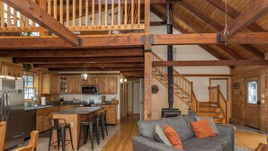 A spacious kitchen with wooden beams, a dining area with bar stools, a gray sofa with orange pillows, and a wooden staircase.