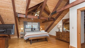 A bedroom with a wooden ceiling, a bed with striped bedding, a bench, a dresser, and a television.