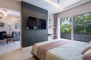 A modern bedroom with a large bed, a TV mounted on a dark wall, a balcony with a view of trees, and a desk with a chair.