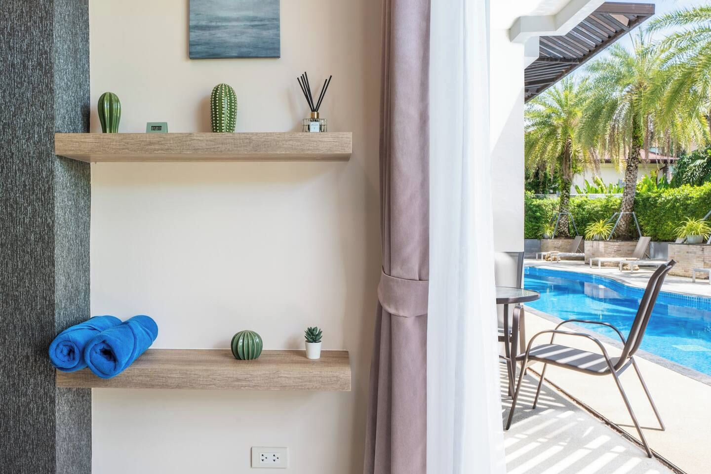 A room with two wooden shelves, a blue towel, a small plant, and a view of a swimming pool with lounge chairs.