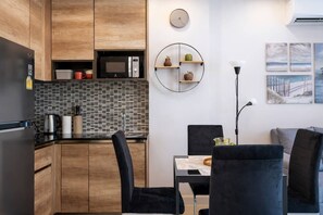A modern kitchen with wooden cabinets, a microwave, and a tiled backsplash. There is a dining area with a table and chairs, and artwork on the wall. - Condo Vireo | Pool View | Rawai (Tambon Rawai)