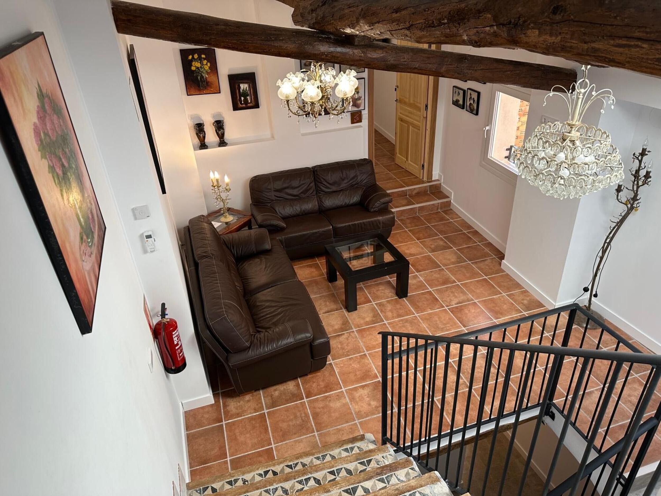 A living room with a brown leather sofa, a glass coffee table, a chandelier, and a staircase with a metal railing.