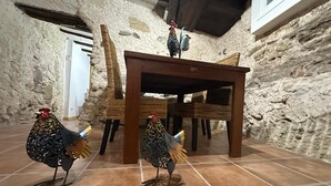 Una habitación rústica con paredes de piedra, una mesa de madera y dos esculturas ornamentales de gallo de metal.