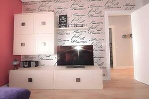 A modern living room with a flat-screen TV mounted on a white cabinet, a pink wall, and a wallpaper with repeated text.