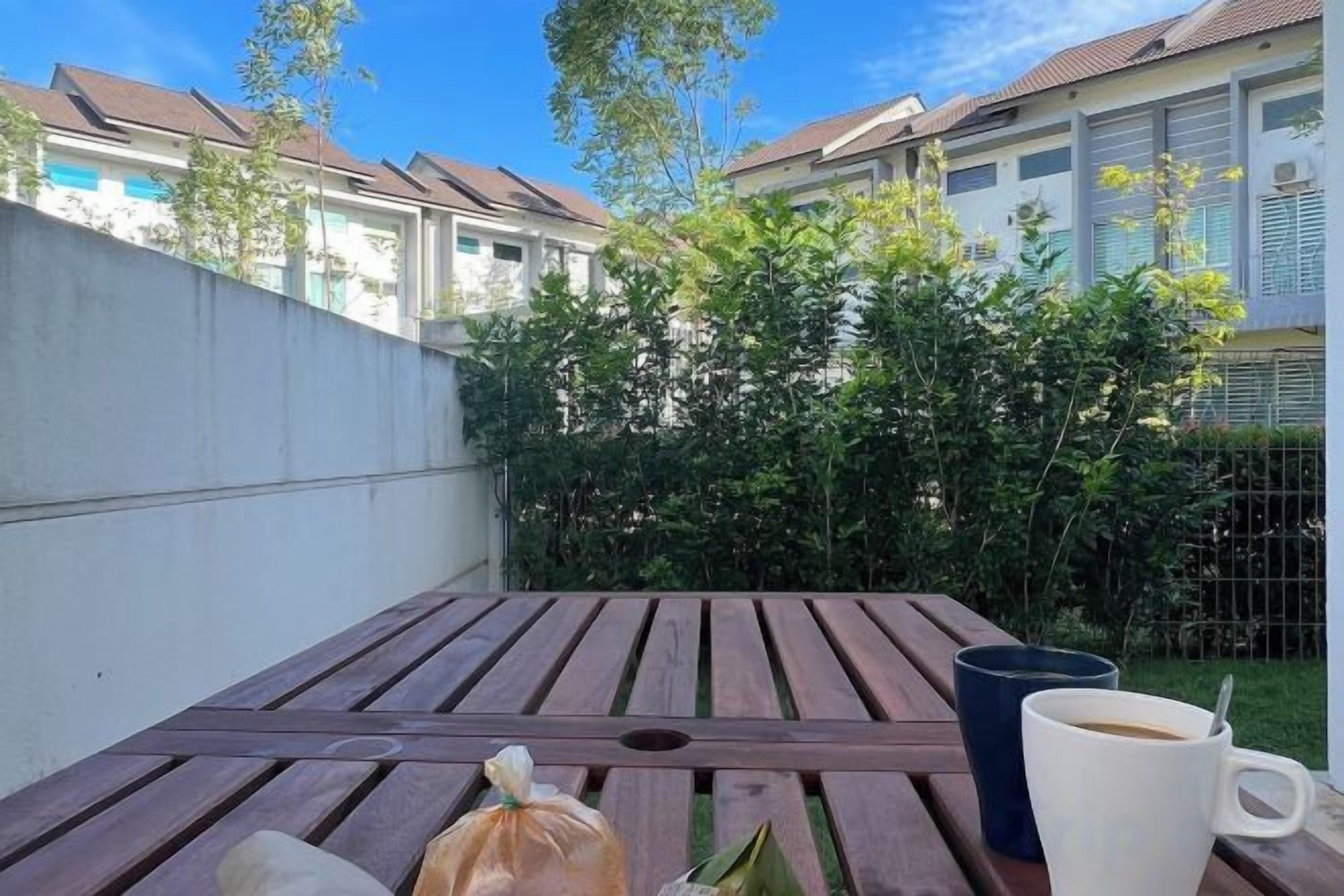 A wooden table with a bag and a mug, set against a backdrop of a white wall and a fence with greenery.