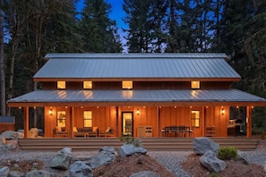 Une cabane en bois à deux étages, avec un toit en métal, de grandes fenêtres et une véranda couverte équipée de sièges.