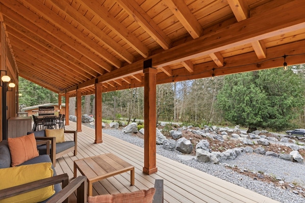 Une terrasse extérieure couverte, aménagée avec du mobilier en bois, un barbecue et offrant une vue sur une zone boisée.