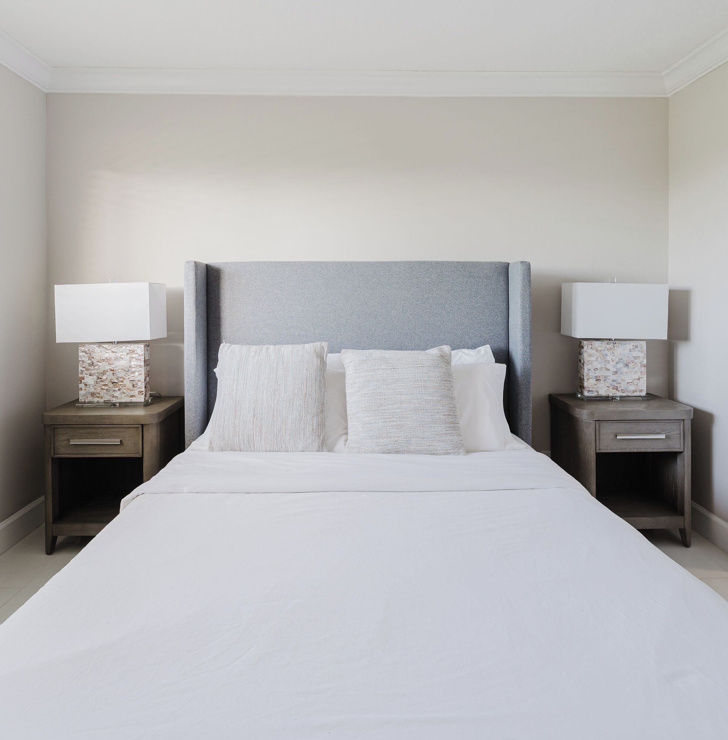 A neatly made bed with a grey headboard, two bedside tables with lamps, and a plain wall.