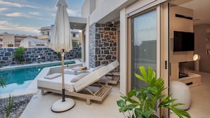 A modern outdoor pool area with lounge chairs and a large umbrella, surrounded by stone walls and a glass door leading to a living area.