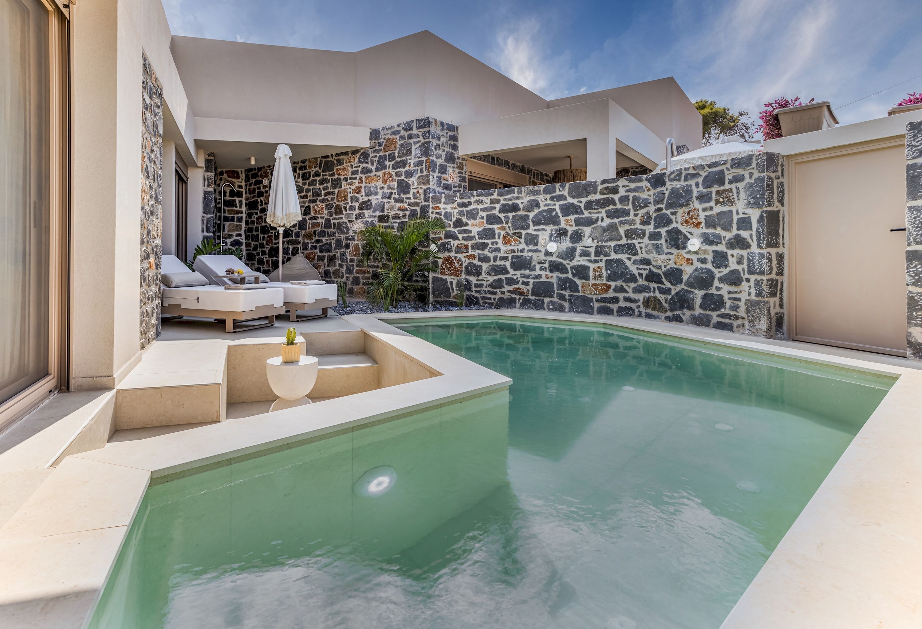 A modern outdoor pool area with stone walls, lounge chairs, and a small table.