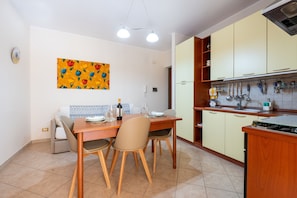 Een eethoek met een tafel en stoelen, een keuken met kastjes en keukengerei, en een kleurrijk schilderij aan de muur.
