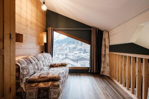A cozy room with a wooden sofa, a large window offering a view of a snowy village, and a wooden railing.